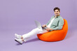 Full length fun smiling man 20s wear casual mint shirt white t-shirt sitting in orange bean bag chair use laptop pc computer chat isolated on purple color background studio People lifestyle concept