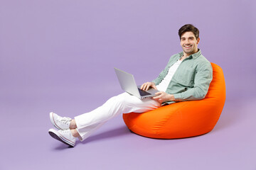 Wall Mural - Full length fun smiling man 20s wear casual mint shirt white t-shirt sitting in orange bean bag chair use laptop pc computer chat isolated on purple color background studio People lifestyle concept