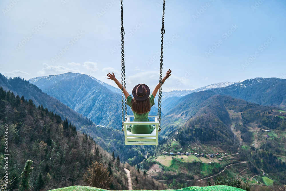 Free happy joyful woman traveler with open arms swinging on chain swing in the mountains, enjoying beautiful view and good life moment - obrazy, fototapety, plakaty 