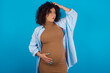 © Jihan - young Arab pregnant woman wearing dress against blue wall confuse and wonder about question. Uncertain with doubt, thinking with hand on head. Pensive concept.