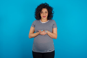 Canvas Print - Photo of cheerful confident young Arab pregnant woman wearing stripped t-shirt standing against blue background arms together