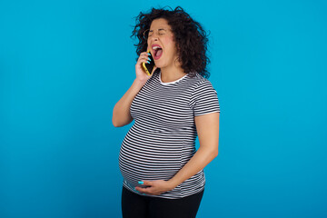 Sticker - Overemotive happy young Arab pregnant woman wearing stripped t-shirt standing against blue background laughs out positively hears funny story from friend during telephone conversation