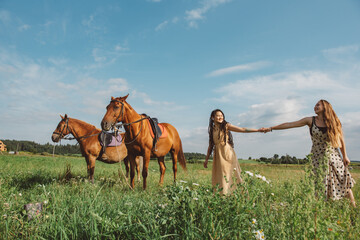 Two young beautiful girls in long summer dresses are walking in the field with two horses.Girls are resting in nature on a sunny summer day while riding horses.Clear blue sky.Defocus, selective focus.