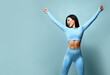 Portrait of fitness girl. Woman in fashion sportswear, standing on a blue background, after a workout. Fit girls with a strong muscular body, raised her arms up, rejoices in her body