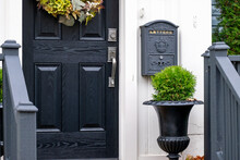 A Closed Solid Wood Black Front Door Of A House. There's A Black Mailbox Hanging On The White Exterior Wall. There's A Flower Pot With A Green Hedge. The Door Has A Colorful Flower Wreath. 