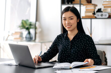 Portrait Of A Pretty Asian Young Woman. Successful Confident Japanese Female Office Worker, Wearing Formal Shirt, Sitting At Her Workplace With Laptop, Looking At The Camera, Smiling Friendly
