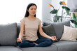 © Kateryna - Calm beautiful young asian brunette woman, in casual clothes, sitting at home on the sofa in the lotus position, resting, taking care of her health, relaxing and meditating, closing her eyes