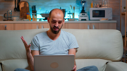 Poster - Caucasian male sitting on couch in pajamas working on laptop computer during videocall conference. Comfortable man relaxing on sofa chatting with business people late at night in kitchen