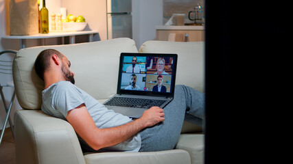Poster - Tired man in pajamas sitting on sofa falling asleep while having online business videocall with teammates using laptop computer. Caucasian male on conference internet web communication