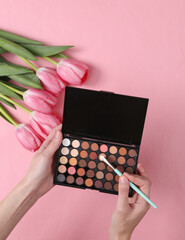 Wall Mural - Hands holding Make-up eye shadow palette with brush and Pink tulips on pink background. Romantic, beauty, love concept