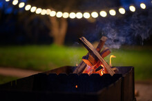 Wooden Logs Burning In The Grill In The Night Garden Decorated With Lanterns. Summer Parties With Grill Cocept