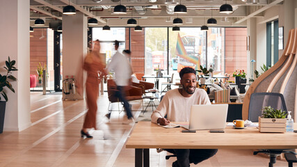 Businessman Sitting At Desk Writing In Notebook In Modern Open Plan Office