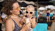 © Sia - Young Caucasian mother holds her baby girl drinking a milkshake from glass with a paper straw. Kids friendly beach party