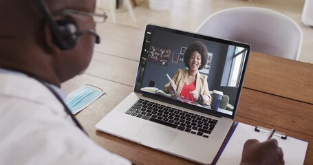 Wall Mural - African american male doctor wearing phone headset taking notes while having a video call on laptop