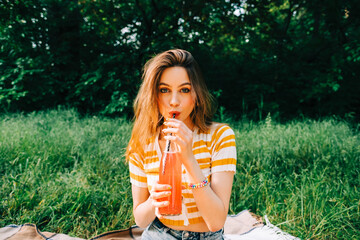 Wall Mural - Young cheerful woman with drink, in natural park on picnic, enjoying summer holidays vacation.