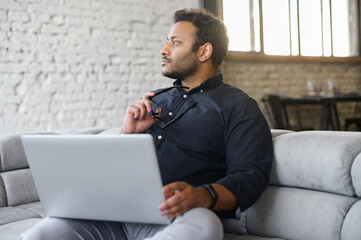 Wall Mural - Contemplative mixed-race hindu guy sits on the sofa with a laptop and waiting for inspiration, looks away, figured out how to solve an important problem or project issues