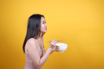 Wall Mural - Beautiful young south east Asian woman holding chines empty chopstick Chinese soup spoon bowl utensil pretend acting posing see eat taste smell feed offer satisfaction yummy yellow orange background