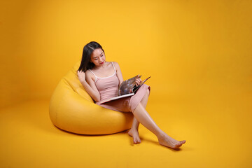 Wall Mural - Beautiful young south east Asian woman sit on a yellow orange beanbag seat color background relax read study book magazine