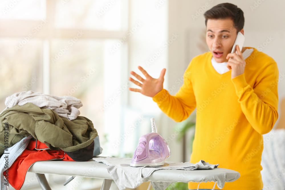 Shocked young man with laundry talking by mobile phone at home