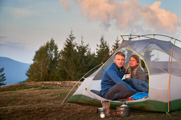 Wall Mural - Portrait of pair of hikers sitting in their tent after breakfast outdoors in the mountains. Green forest, mountain hill and morning sky on the background. Concept of hiking, camping and relationships.