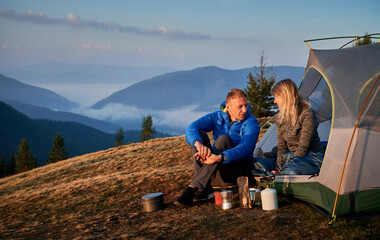 Wall Mural - Two travelers happily talking about their vacation in the mountains, resting near tent, laying out special equipment for cooking outdoors. On the background beautiful view of the mountain hills.