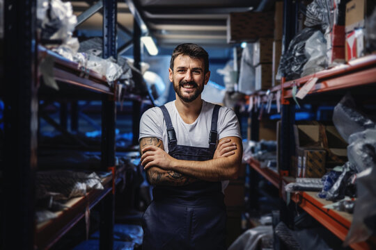 Young smiling happy tattooed bearded worker in overalls standing in storage of import and export firm with arms crossed.