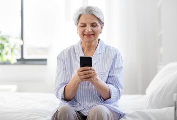 Canvas Print - old age, technology and people concept - happy smiling senior woman in pajamas with smartphone sitting on bed at home bedroom