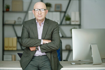 Portrait of serious mature Caucasian businessman in formal suit standing with crossed arms against office table with modern computer