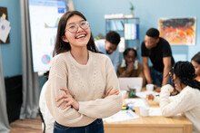 Portrait Of An Asian Woman Standing With Arms Crossed Over Her Chest, Wearing Sweater And Glasses She Smiles. In Background View Office, Interactive Whiteboard, Coworkers Working On Project At Table