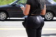 Woman with big buttocks in tight pants stands by the road with smartphone in her hand on car background. Female fashion, car sharing, calling a taxi concept