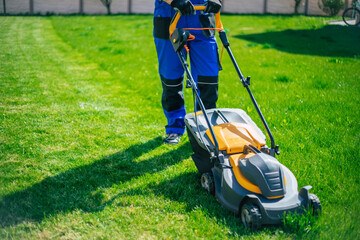 Young man mows the lawn using an electric lawn mower in a special worker suit near a large country house in the backyard