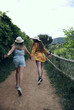 © Gusta Cabrera - Two girls with hats smiling and running backwards along a path with many plants.