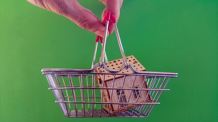 Real estate market and real estate purchase, the fingers of a person holding a house icon and a shopping cart. Buying a home concept on a green background. Copy space.