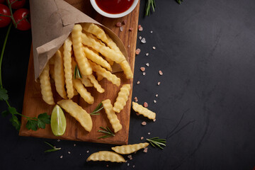 Wall Mural - Homemade baked potato fries with mayonnaise, tomato sauce and rosemary on wooden board. tasty french fries on cutting board, in brown paper on black stone table background, unhealthy food.