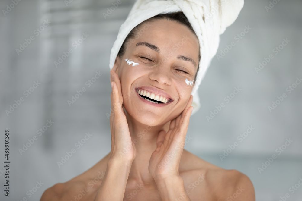 Happy excited beautiful girl enjoying facial skincare procedures, applying nourishing cream for smooth silky eye skin, massaging face with fingers, with closed eyes, smiling, laughing, Head shot - obrazy, fototapety, plakaty 