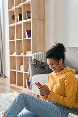 Vertical shot of happy smiling young African American mixed raced girl holding smartphone sitting on floor at home using online entertainment mobile applications watching streaming, shopping online.