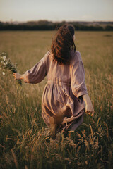 Wall Mural - Beautiful woman in linen dress running with wildflowers in hand in summer meadow in sunset. Stylish young female in rustic dress enjoying free evening in countryside. Atmospheric carefree moment