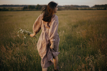 Wall Mural - Beautiful woman in linen dress running with wildflowers in hand in summer meadow in sunset. Atmospheric carefree moment. Stylish young female in rustic dress enjoying free evening in countryside