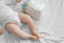 Baby Feet And A Pack Of Diapers On A White Bed. Dirty Diapers. Newborn And Costs