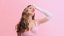 A Beautiful Young Woman Wearing Gorgeous Evening Pink Dress With Gloves And Hairpins Is Losing Consciousness Isolated Over A Pink Wall In The Studio