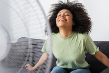 Pleased African American Woman Sitting On Couch Near Blurred Electric Fan