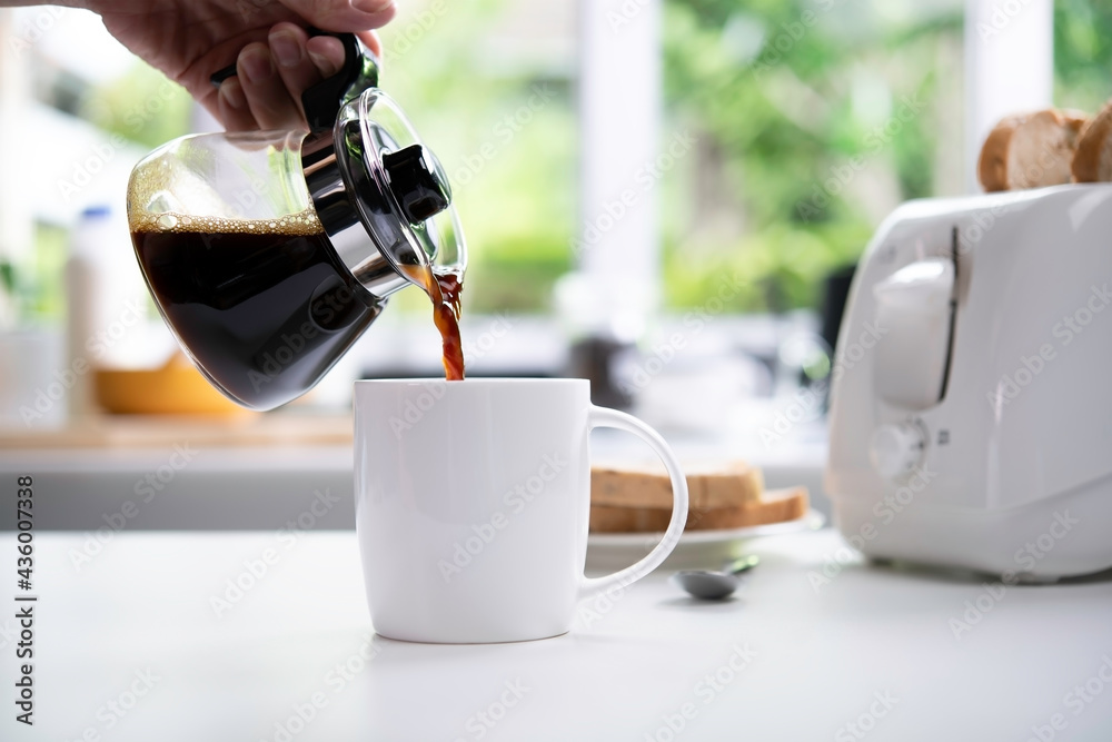 A hand pouring steaming coffee in to a cup on table in the kitchen - obrazy, fototapety, plakaty 