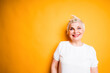 © DmitryStock - Cheerful beautiful elderly woman in a white t-shirt stands on a yellow background and smiles while looking at the camera