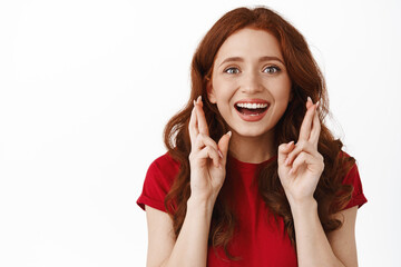 Wall Mural - Close up portrait of hopeful ginger girl, cross fingers for good luck, looking happy and smiling, anticipating something, waiting for exciting results, standing against white background