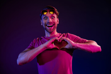 Sticker - Portrait of attractive amazed cheerful guy showing heart shape having fun isolated over dark neon purple color background