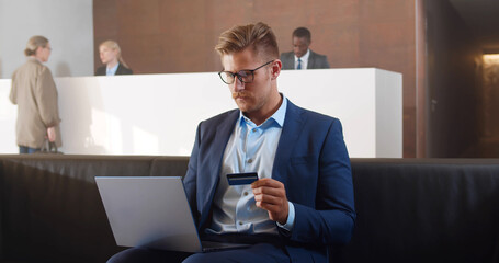 Handsome young businessman wearing suit sitting at hotel lobby using laptop and credit card