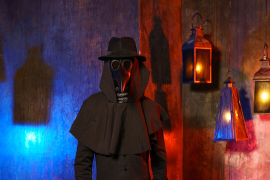 Portrait of a plague doctor in a black leather mask, hat and hood, isolated against a dark wall with lanterns. Stylization of a historical costume, a creepy image, the concept of an epidemic.