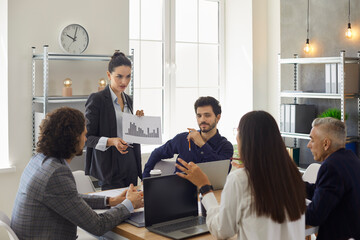 Wall Mural - Strict team manager standing at office table in corporate group meeting, holding column chart paper, showing sales increase decrease figures and listening to female coworker making smart suggestion