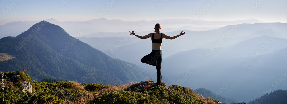 Back view of woman practicing yoga in evening mountains. Meditating female is balancing on one leg after sunset outdoor. Concept of harmony with nature. - obrazy, fototapety, plakaty 