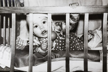 Little Baby In Bed Looking Through The Wooden Rods. Vintage Black And White Paper Photo. Early 1980s. Old Surface, Soft Focus. Transferred Property, Family Archive.
